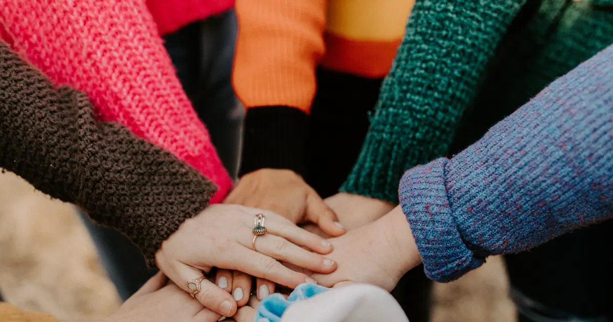 Arms and hands of women wearing colorful sweaters in a circle with hands piled together.