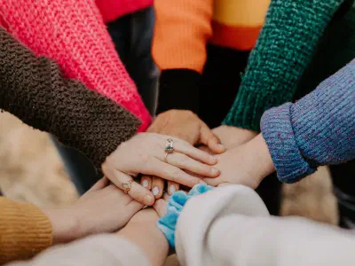 Arms and hands of women wearing colorful sweaters in a circle with hands piled together.