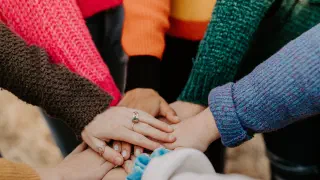 Arms and hands of women wearing colorful sweaters in a circle with hands piled together.